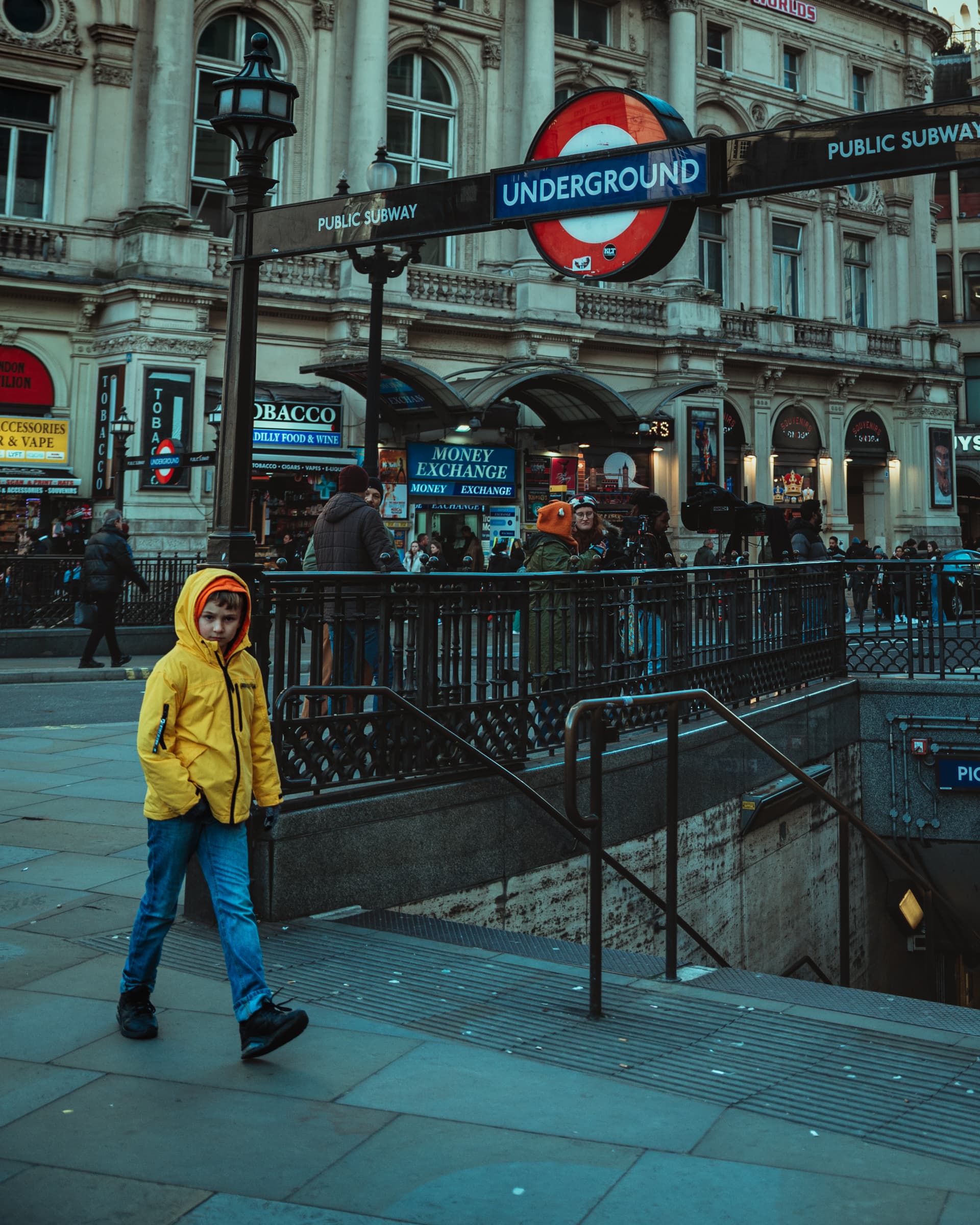 Child in a yellow raincoat walking past a London Underground entrance