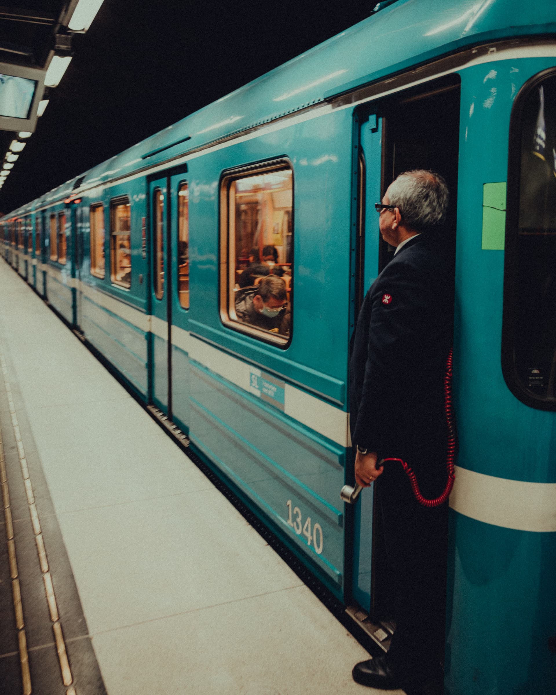 Conductor in uniform leaning out of a teal train at a platform