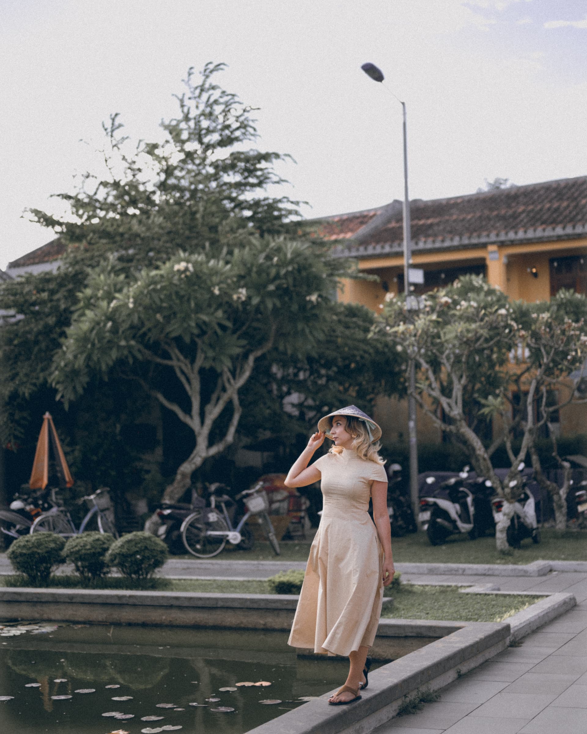 Woman in a sun hat and cream dress by a pond in front of a yellow colonial building
